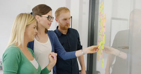 Obraz premium Side view of two women and male coworker creating new ideas on project and putting stickers on glass wall of office.
