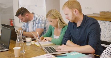 Young concentrated people writing in documents and using laptop while sitting at wooden table in office.