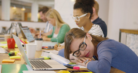 Woman in glasses lying on table in office and sleeping on background of young colleagues working with documents.