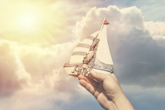 Handmade ship in a man's hand against a background of clouds as a symbol of travel and dreams