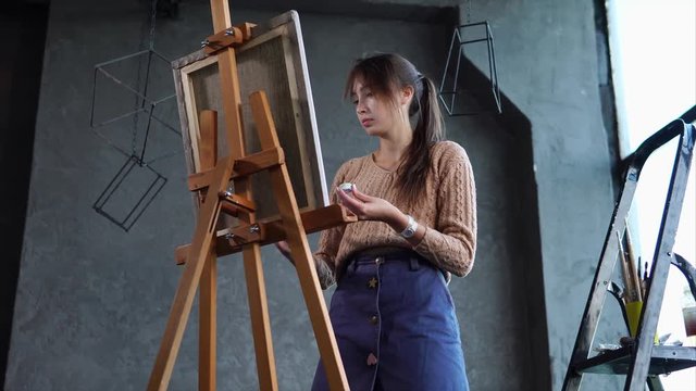 Quite a skilfull artist is drawing a picture on an easel in art studio with black walls. She is painting with a brush very fast.