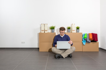 Mann sitzt auf dem Boden im leeren Raum. Er bedient ein Notebook und schaut aufs Display