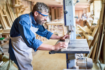 Side view portrait of mature bearded carpenter carefully placing piece of wood on cutting machine in modern workshop