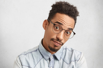 Serious surprised male with oval face, specific appearance, looks through big spectacles, wonders why work hasn`t done yet. Young male adult wears denim shirt, expresses wonderment and amazement