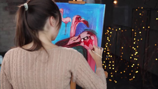 Woman is applying colors on her picture. Camera is filming through her back, showing amazing vivid composition, standing on an easel in workshop.
