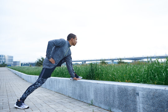 Candid lifestyle shot of muscular strong African American male in trendy outfit stretching legs outdoors, warming up mucles before cardio training. Virality, physical exercise, workout and motivation