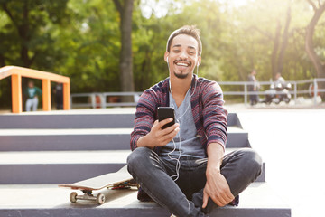 Joyful restful mixed race teenager sits on ground outdoors, uses mobile phone as listens melodies with earphones. Young talented skater rests after long training, enjoys music and sunny weather