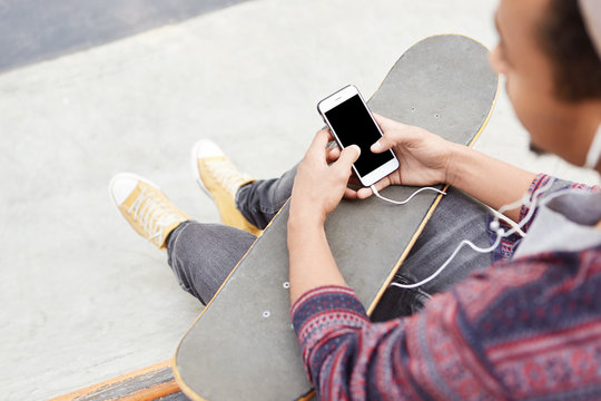 Cropped shot of male teenger has active lifestyle, skateboards on skate play, recieves message from friend, invites him to go in for extreme sport together, listens radio online with earphones