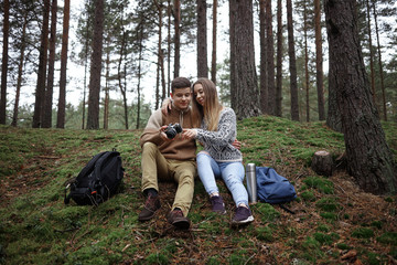 Handsome guy and beautiful girl wearing sensible clothes watching pictures on professional camera, sitting on grass close to each other among trees with backpacks and thermos bottle on the ground