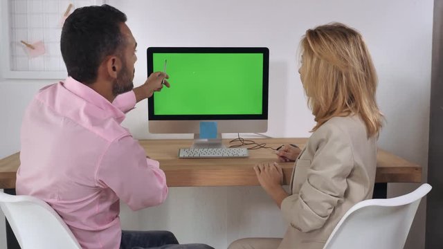 Business people sitting at the desk in front of computer with green screen have conversation. Meeting in the office with white wall and casual interior