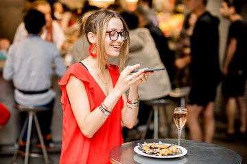 Young woman in red dress photographing plate of mussels with glass of wine sitting at the food market