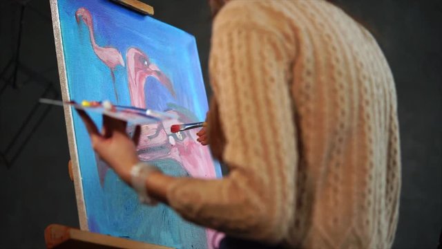 A young woman who draws pink flamingos in different angles stands in the art studio, in her hand is a palette with oil paints, the lady holds a brush