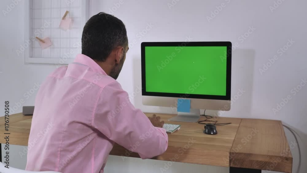 Aufkleber Back view focused businessman wearing casual shirt sitting at the working place works on computer with green display. Man entering data typing on keyboard in home office looking on pc