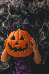 Naklejka premium Little girl dressed as a witch holding a pumpkin against a dark background with spiderwebs