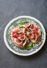 Delicious appetizers for wine or a snack - sandwich with goat cheese, prosciutto and figs on a vintage plate. On a gray background, top view