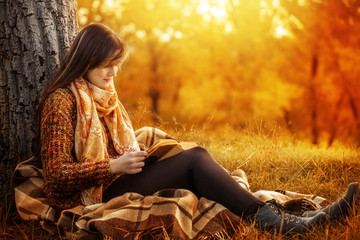 Beautiful young brunette sitting on a fallen autumn leaves in a park, reading a book