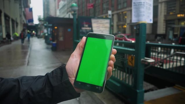 A man holds a green screen smartphone outside of a Manhattan subway entrance on a rainy day. With optional corner pin markers for advanced screen replacement.  	