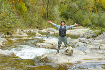 An old traveler relaxes on a rocky river, lifting his arms and breathing fresh air. Senior tourist stands on the rocks of the mountain river
