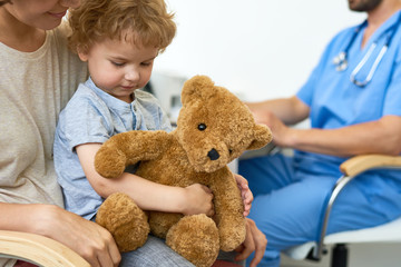 Portrait of cute little child sitting on mothers lap in doctors office waiting for check up hugging plush teddy bear toy