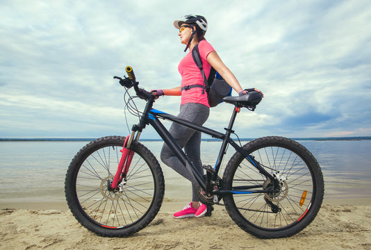 One young woman, an athlete in pink shoes, walks with a mountain bike in the morning on the river bank.