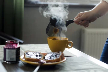  woman pours hot coffee from cezve in  mug in front of the window © dvulikaia