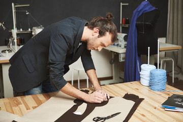 Portrait of manly good-looking adult male clothes designer with stylish hairstyle in black suit cutting out parts of future dress from fabric. Man concentrated on work.