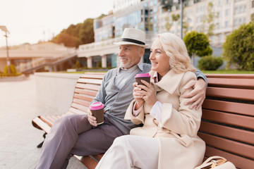 Two pensioners are sitting on a bench with a glass of coffee in their hands. They sit embracing and smiling.