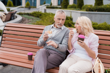 Fototapeta premium An elderly couple is resting sitting on a bench in the square. They eat burgers and drink coffee