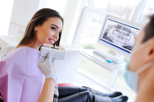 The dentist talks with the patient about dental care and selects the patient the color of the tooth enamel against the background of the monitor with the X-ray of the jaw