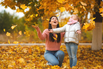Cheerful mother and her little daughter having fun together in the autumn background. Happy family in the fall background. Cute girls with colorful leaves.