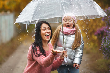 Cheerful mother and her little daughter having fun together in the autumn background under the umbrella. Happy family in the fall background. Cute girls with umbrella.