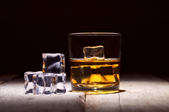 Glass of whiskey with ice cubes served on white wooden planks.