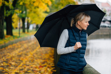 Pretty young blonde smiling woman in waistcoat with umbrella standing in colorful autumn park.