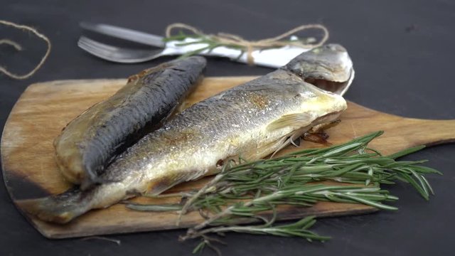 Baked fish on a cutting board, next to it there is a rosemary, in the background there are cutlery in a napkin bandaged with string