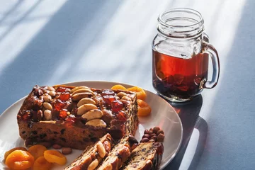 Foto op Canvas All butter cake packed with fruits and topped with glazed cherries and mixed nuts. Accompanied with a cup of black tea, dried apricots and mixed nuts. Light blue background © Mister Dreamer