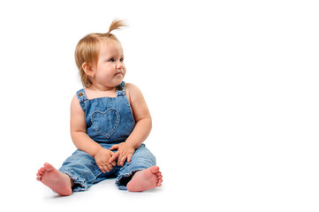 Cute little baby girl with funny haircut in jeans overalls sitting on white background and looking to the side, isolated