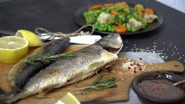 Fried fish on a cutting board around scattered rosemary, lemon, sesame and red berries on a plate next side is a garnish of vegetables