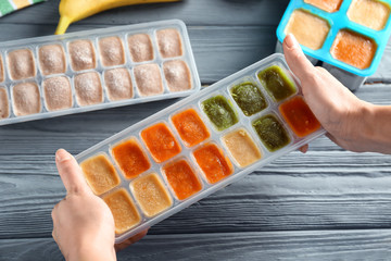 Woman holding ice tray with frozen vegetable puree near table