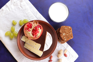 Camembert cheese, a glass of kefir, pomegranate, berries of green grapes, peanut grains and bread on white and dark blue background