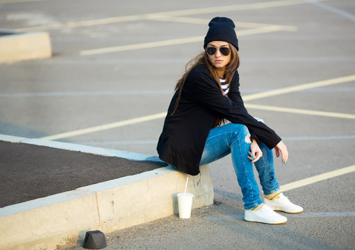 Outdoor fashion closeup portrait of nice pretty young hipster woman posing in sneakers and sunglasses sitting on the curb with a drink at sunset.