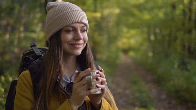 Happy female tourist, in yellow jacket and warm hat, having hot tea while standing in the wood, slowmotion on fall day