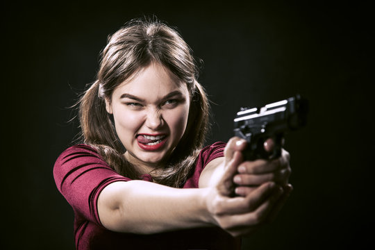 fun girl with gun on black background aiming at camera, show tongue