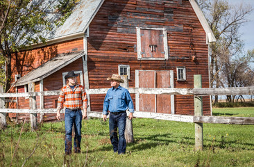 horizontal image of two caucasian cowboys walking and talking next to a wooden fence with a big red barn in the background on a ranch on a sunny summer day.