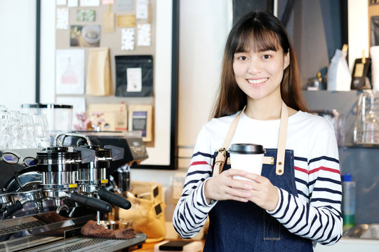 Young asian women Barista holding a take away coffee cup with smiling face at cafe counter background, small business owner, food and drink industry concept