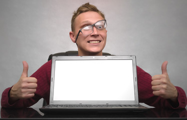 A successful smiling young business man in glasses showing thumbs up sign symbol behind blank screen of laptop computer on the table. Special offer. Big sale. Contact us. Winner. Lucky.