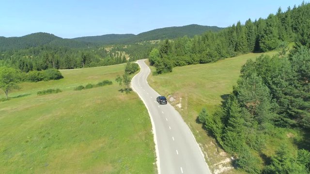 AERIAL: Black SUV car driving along empty country highway in idyllic rural landscape. People on road trip journey across beautiful green Slovenia. Lush green hills and meadow fields in remote valley