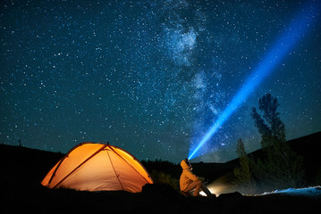 Man tourist with flashlight near his camp tent at night. - obrazy, fototapety, plakaty