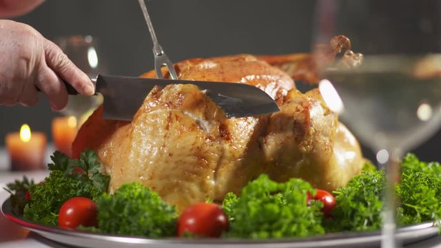 Thanksgiving or Christmas turkey dinner. Carving the turkey in slow motion cutting juicy breast meat. Whole roasted turkey steaming on tray garnished with tomatoes and herbs.