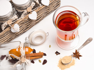 Tea with spices in a glass mug on a white background. Next basket with jars and spices: cinnamon, cardamom, orange, cloves. Spoon with ginger.