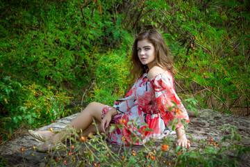 Portrait of beautiful young smiling brunette woman wearing colorful dress, sitting on a stone cliff in a forest on a hot summer day